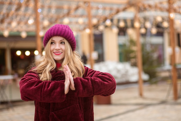 Lovely blonde woman wearing red knitted hat and winter coat, posing on the background of garland at the street in Kyiv