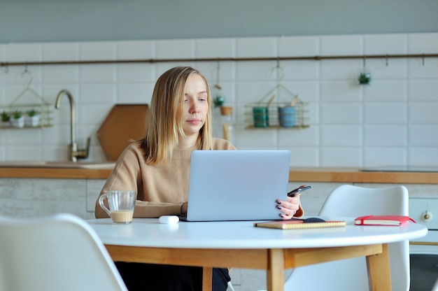 Lovely blonde woman working with her laptop while sitting in the kitchen in her apartment