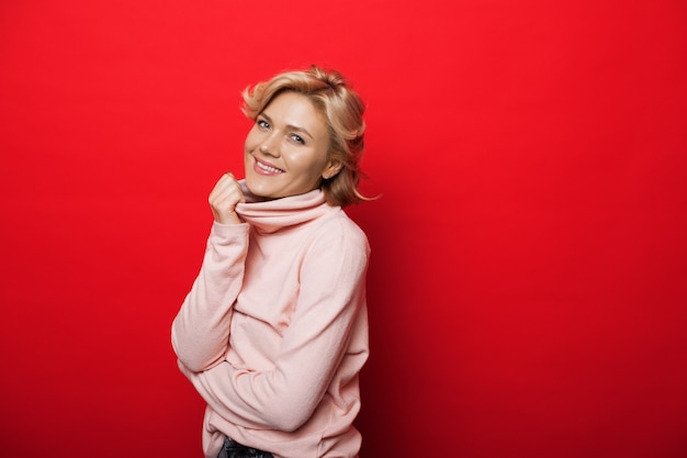 Lovely caucasian woman with blonde hair wearing a pink sweater is smiling at camera on a red studio wall