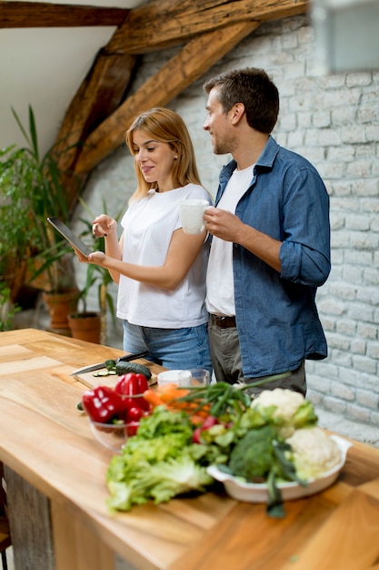 Lovely cheerful couple cooking dinner together, looking recipe at digital tablet  and having fun at rustic kitchen