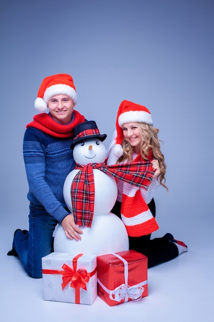 Lovely christmas couple  in Santa Claus hats sitting with presents and snowman on blue background