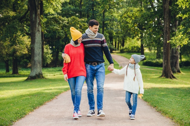 Lovely couple embraces each other, looks at their small good looking daughter, walks across green park