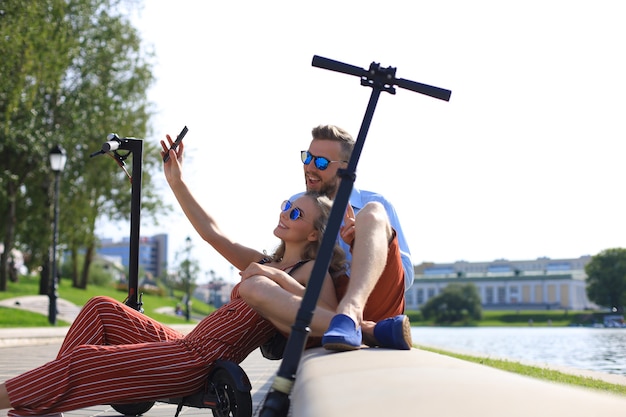 Photo lovely couple having fun driving electric scooter, taking a break from driving, sitting on riverbank, taking selfie,