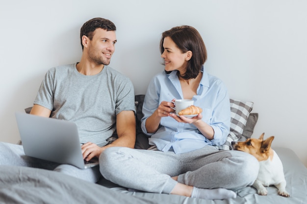 Lovely couple sit together on bed, use laptop computer, have pleasant conversation between each other