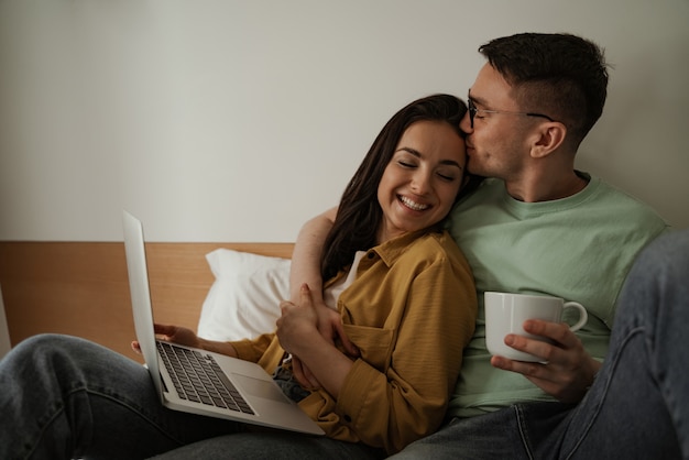 Lovely couple watching movie with romantic feelings, sitting on bed with coffee. Happy family moving to new apartment.