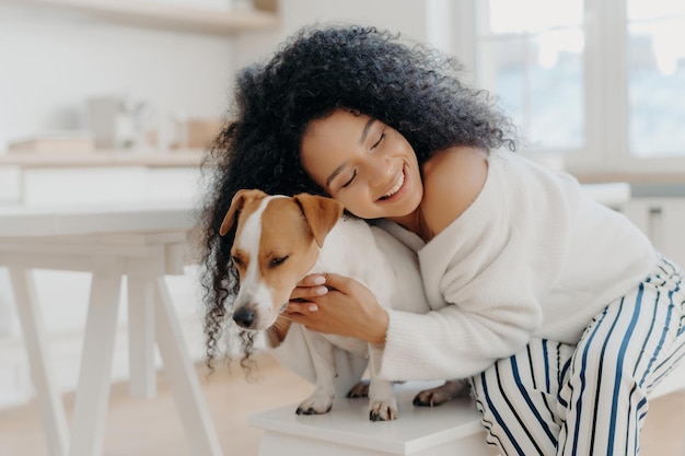 Lovely curly woman embraces beloved dog with care wearing stylish clothes posing at home expressing affection