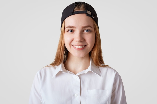 Lovely cute female with positive smile, wears white elegant shirt and stylish cap, going to school or college in good mood. Hipster girl spends time with friends