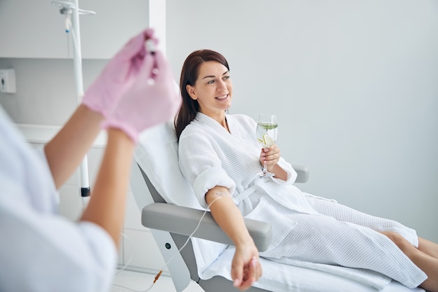 Lovely dark-haired female patient with a glass of drink in her hands undergoing the intravenous vitamin therapy