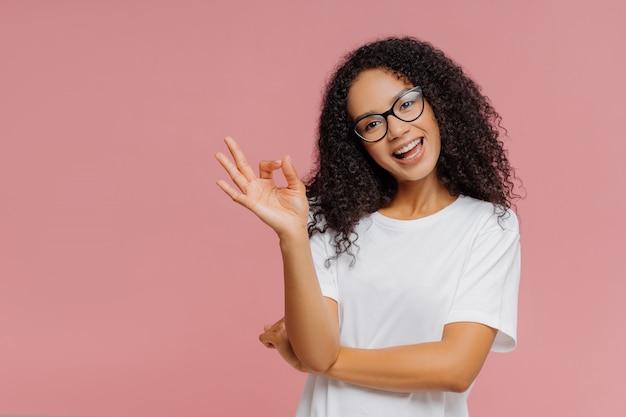 Lovely dark skinned woman makes okay gesture, tilts head, demonstrates approval, agrees with something