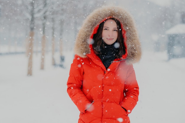 Lovely female model in red jacket stads outdoor during snowfall looks with dark eyes at camera going to have stroll with boyfriend breathes fresh air People winter time and free time concept