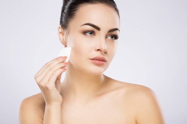 Lovely girl with black hair fixed behind, big eyes, thick eyebrows and naked shoulders standing at gray studio background and holding cosmetic cleaning sponge, looking right.