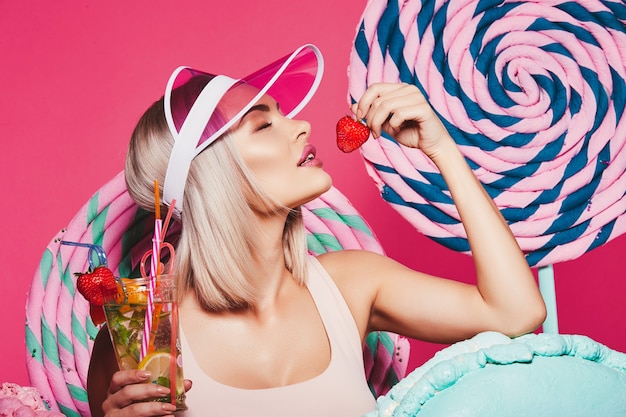 Lovely girl with blonde hair wearing top and pink cap standing with huge sweet lollypops at pink