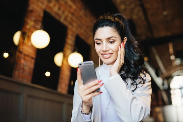 Lovely lady with dark hair makeup and wonderful manicure wearing white jacket holding mobile phone texting SMS listening to music online on earphones enjoying free wireless internet connection