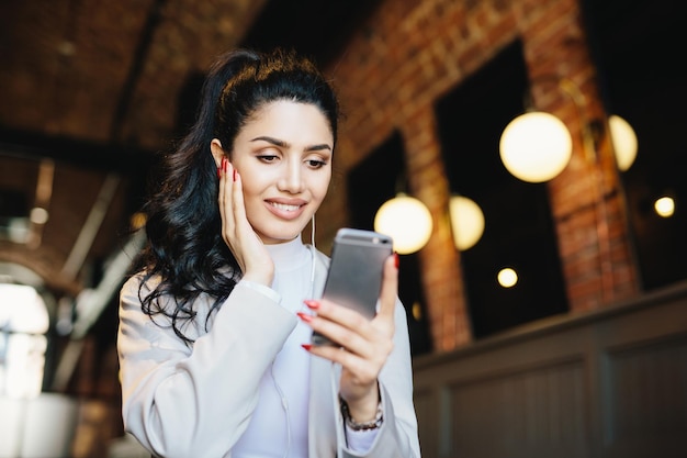 Lovely lady with dark hair makeup and wonderful manicure wearing white jacket holding mobile phone texting SMS listening to music online on earphones enjoying free wireless internet connection