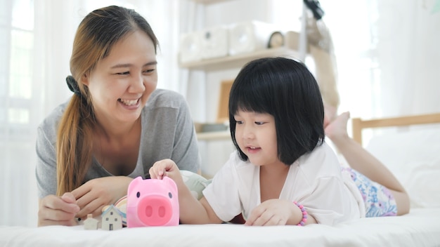 Lovely little Asian girl and mother putting money in piggy bank for saving