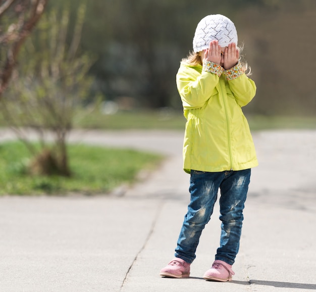 Lovely little girl in yellow coat hiding face with hands