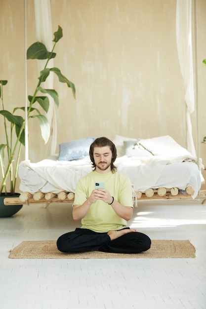 Lovely man sitting in yoga asana using phone, handsome meditation teacher with long hair on yoga mat at home holding smartphone