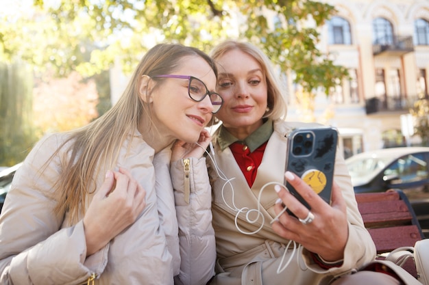 Lovely mature women listening to music together on smart phone, wearing earphones