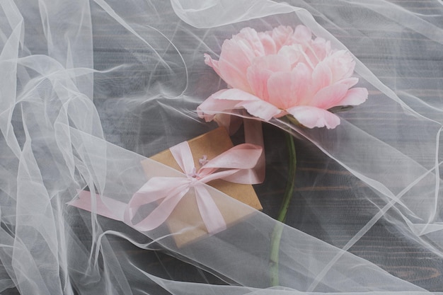 Lovely pink peony flower and gift box under soft tulle fabric on dark wood top view Mothers day