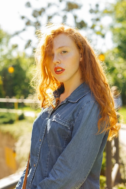 Lovely red haired model in denim shirt posing  in rays of sun at the street