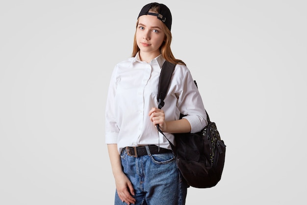 lovely schoolgirl wears jeand and white shirt, carries rucksack, stands on white, goingt to school. Education and learning