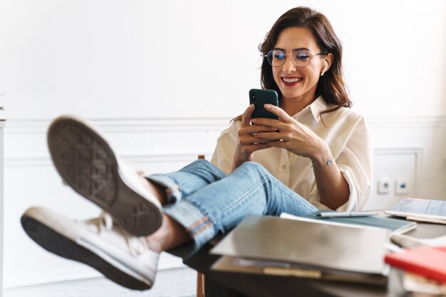 Lovely smiling young brunette woman relaxing at the table in the cafe indoors, using mobile phone