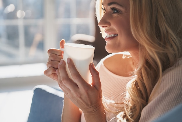 Lovely woman. Attractive young woman holding a cup and looking away with smile while sitting on the sofa at home