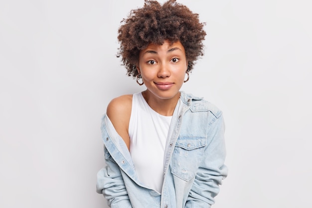 Lovely woman looks with confused expression shows bare shoulder takes off denim jacket has hesitant expression doesnt know what to do isolated over white wall