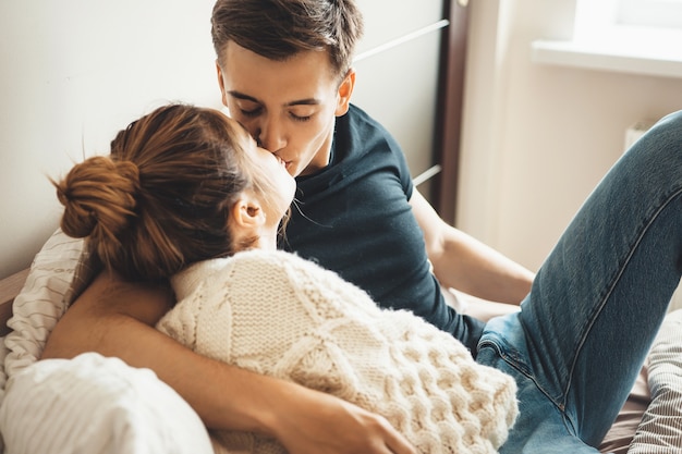 Lovely woman wearing a white knitted sweater kissing her husband while lying in bed on pillows