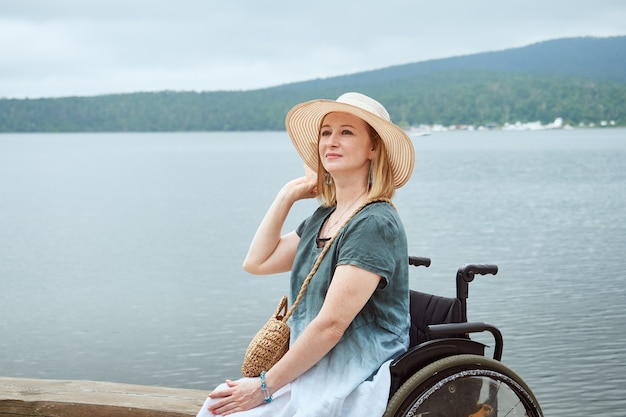 Lovely woman in wheelchair with hat enjoying the seascape or lake.