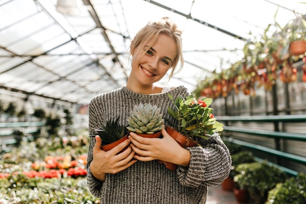 Lovely woman with sweet smile looks into camera holding 3 different plants in brown pots