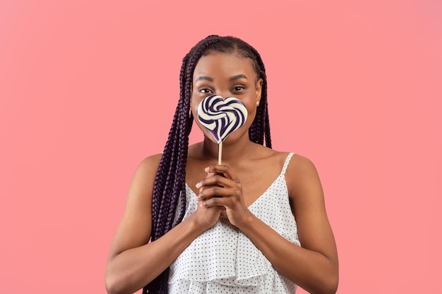 Lovely young black woman covering her mouth with striped heart shaped lollipop on pink studio