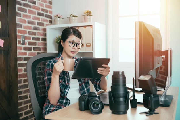 lovely young business photo company worker sitting in office drinking hot coffee and using mobile digital tablet computer checking photographer picture for editing.