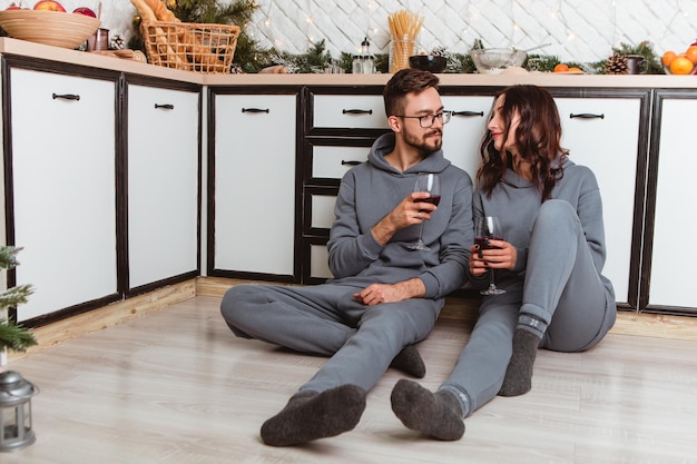 Lovely young couple sitting floor kitchen drink wine in glasses, garlands home cosy interior