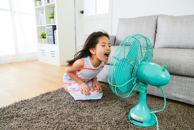 lovely young female children face to electric fan laughing and playing cool wind enjoying summer vacation at home in living room.