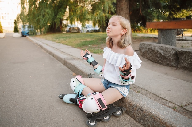 Lovely young girl wearing protective gear and rollerblades sitting on the ground in lotus position, meditating