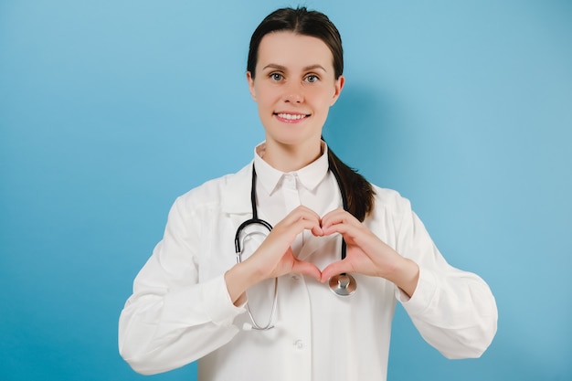 Lovely young woman doctor showing heart gesture and smiling, taking care of patients with love, wears white lab coat stethoscope, models over blue studio background wall. Covid-19, pandemic concept