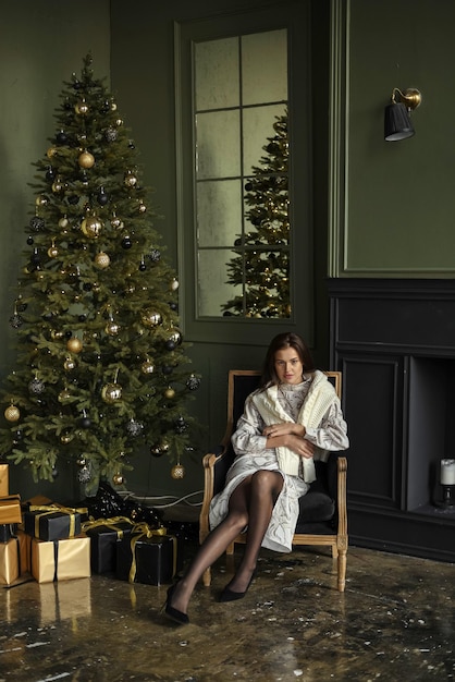 A lovely young woman is warming herself by the fireplace in a sweater near the Christmas tree