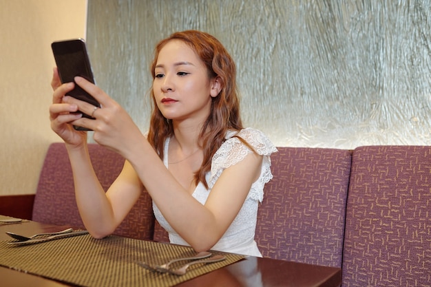 Lovely young woman sitting cafe table and checking application on smartphone when waiting for her order