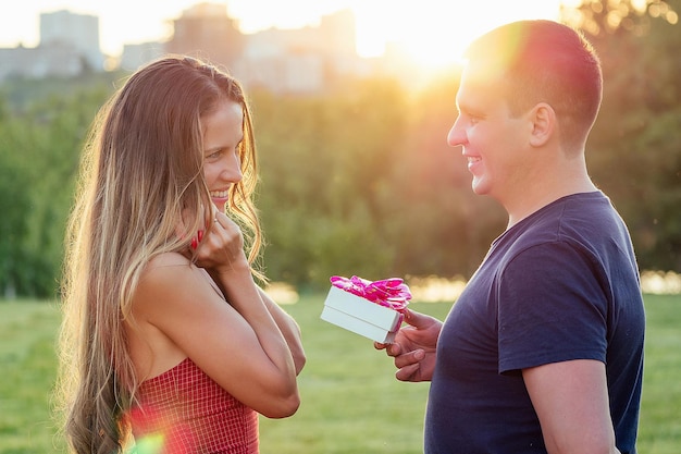 Lover man gives a gift box to embarrassed attractive beautiful blonde long hair woman in love in evening cocktail dress in summer park with grass greens background. first date concept