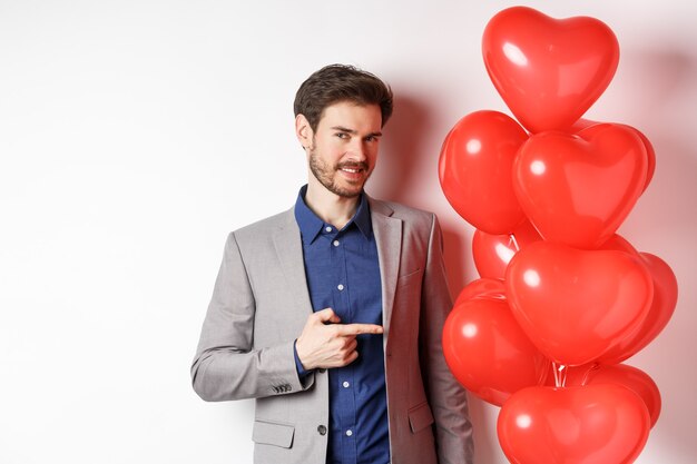 Lovers day. Charming young man with beard, wearing fancy suit, pointing finger at heart balloon surprise for Valentines day, standing over white background.