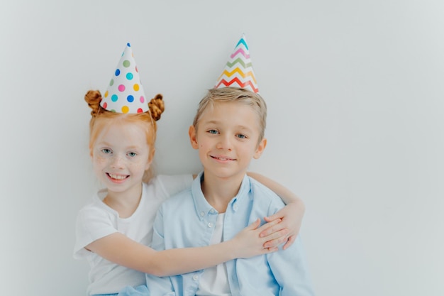 Lovey red haird girl embraces with love her older brother congratulates with birthday wear cone party hats have good mood on party isolated over white background have friendly relationship