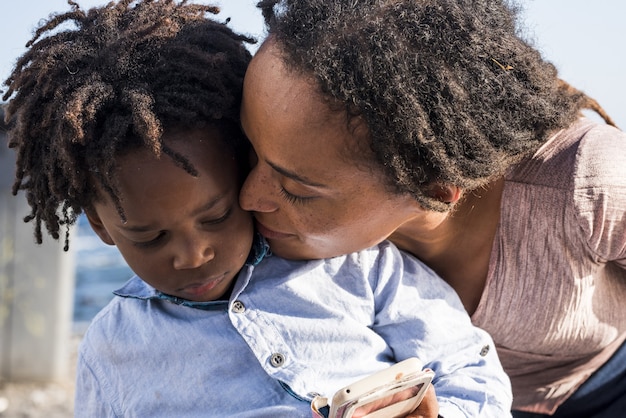 Loving african american mother kissing her little son watching video or playing game using mobile phone outdoors. Caring mother kissing her little son outdoors on a bright sunny day