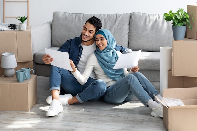 Loving arab family holding papers, working on design for their new house, sitting on floor by sofa among boxes with belongings, holding apartment plan, thinking about home design, copy space