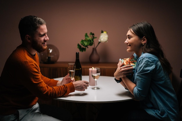 Loving boyfriend giving box with gift to charming happy girlfriend sitting at table with candles on birthday or Valentines Day