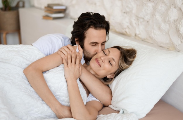 Loving caucasian couple relaxing in bed at home enjoying togetherness and tenderness in love man kissing wife