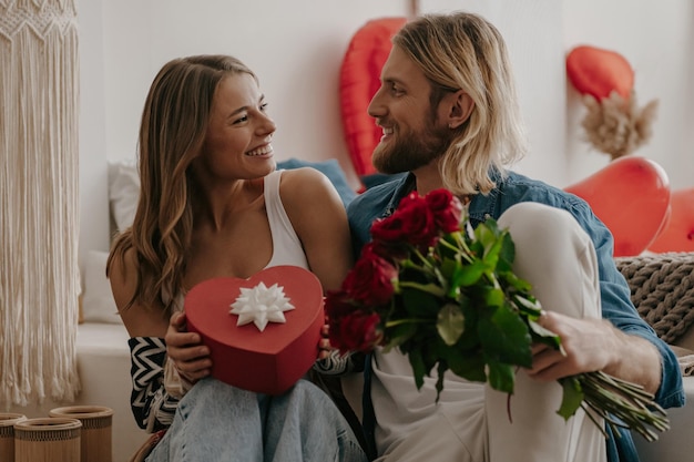 Loving couple embracing and holding roses and gift box while spending valentines day at cozy home