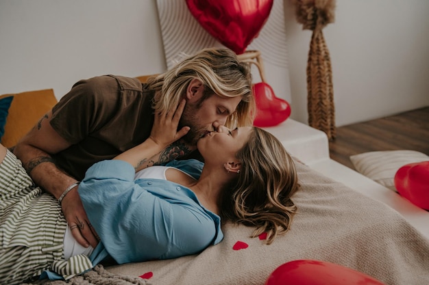 Loving couple embracing and kissing while lying down in bed surrounded with red heart shape balloons