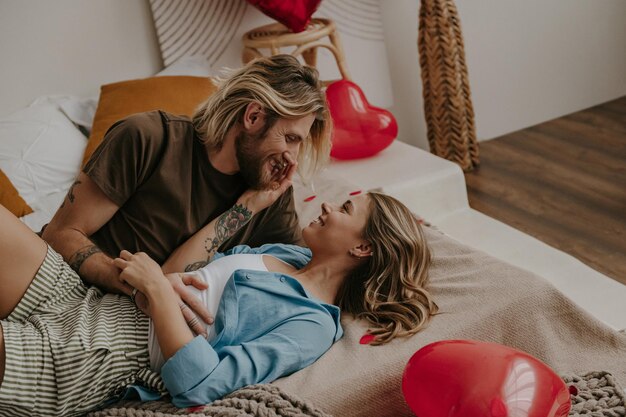 Loving couple embracing and smiling while lying in bed surrounded with red heart shape balloons