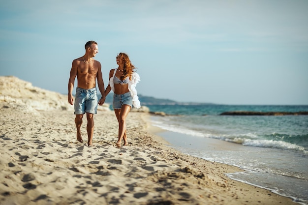 A loving couple is having fun and walking on the empty sandy sea beach.They are looking each other and happily smiling.